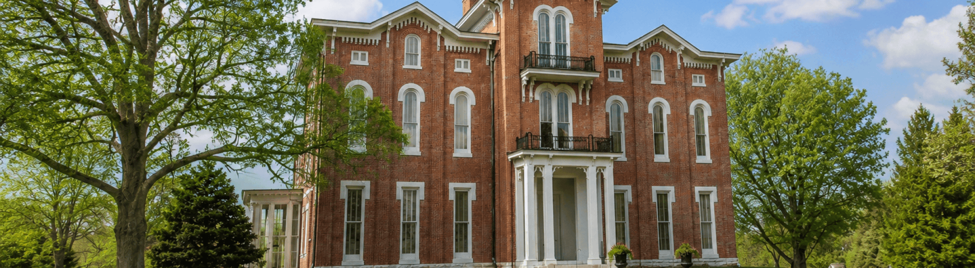 Historic red-brick house with large trees, under a blue sky with clouds.